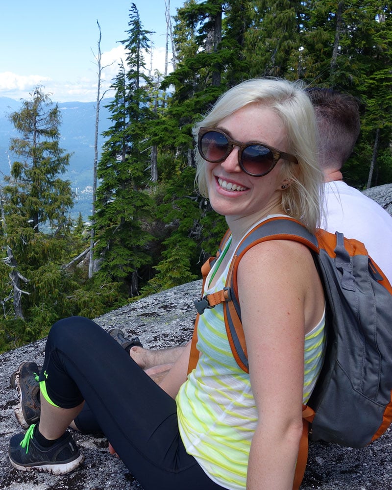 A blonde female in sunglasses and wearing a backpack, sits with a male amongst green pine trees high atop a mountain.